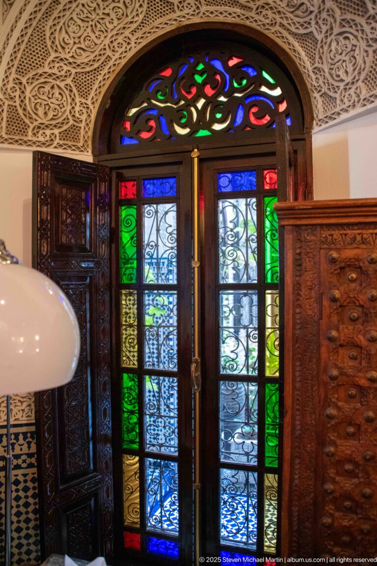 Angled view of carved double doors with colored glass, wrought-iron grilles, fanlight, and carved plaster surround.