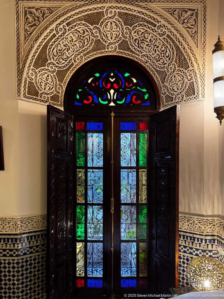 Frontal view of carved double doors with multicolored glass, iron scrollwork, fanlight, carved plaster, and zellij.