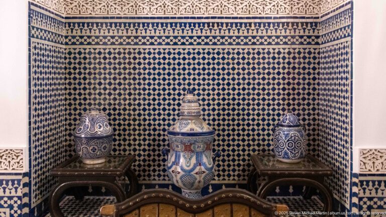 Three painted lidded ceramic vessels on wooden stands in front of a blue-and-white zellij wall.