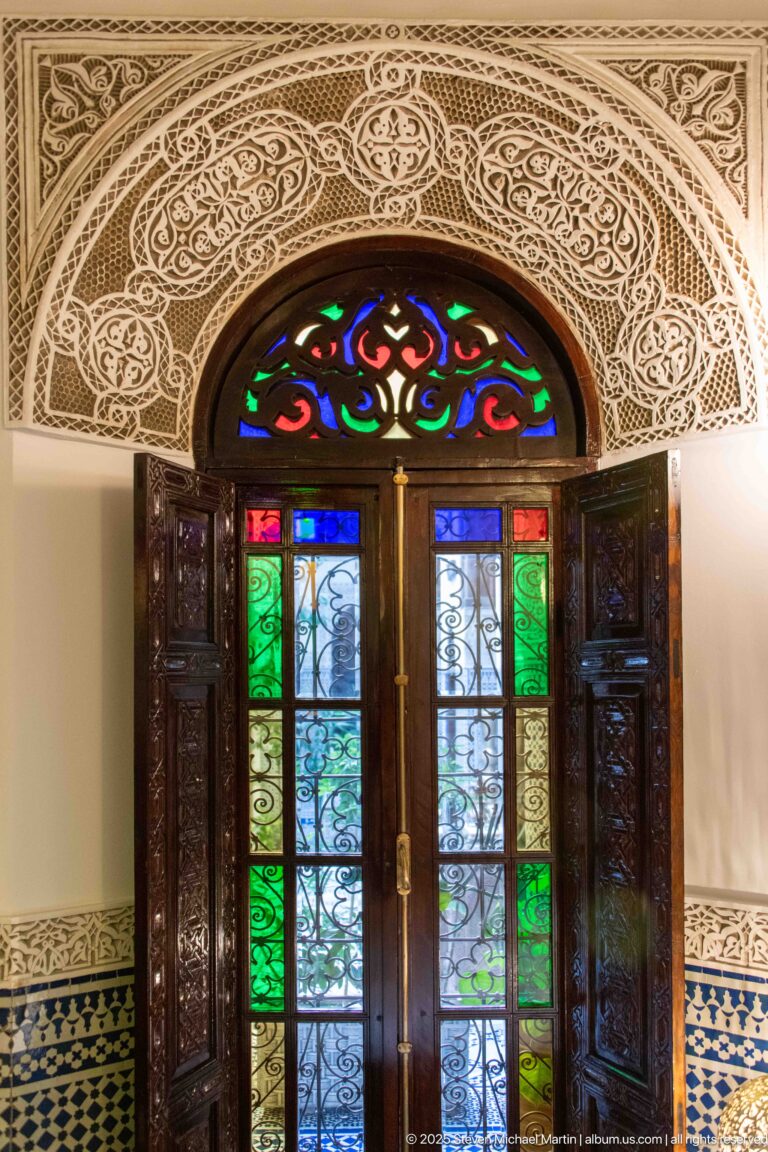 Bright frontal view of carved wooden double doors with colored glass, iron grilles, carved plaster, and zellij.