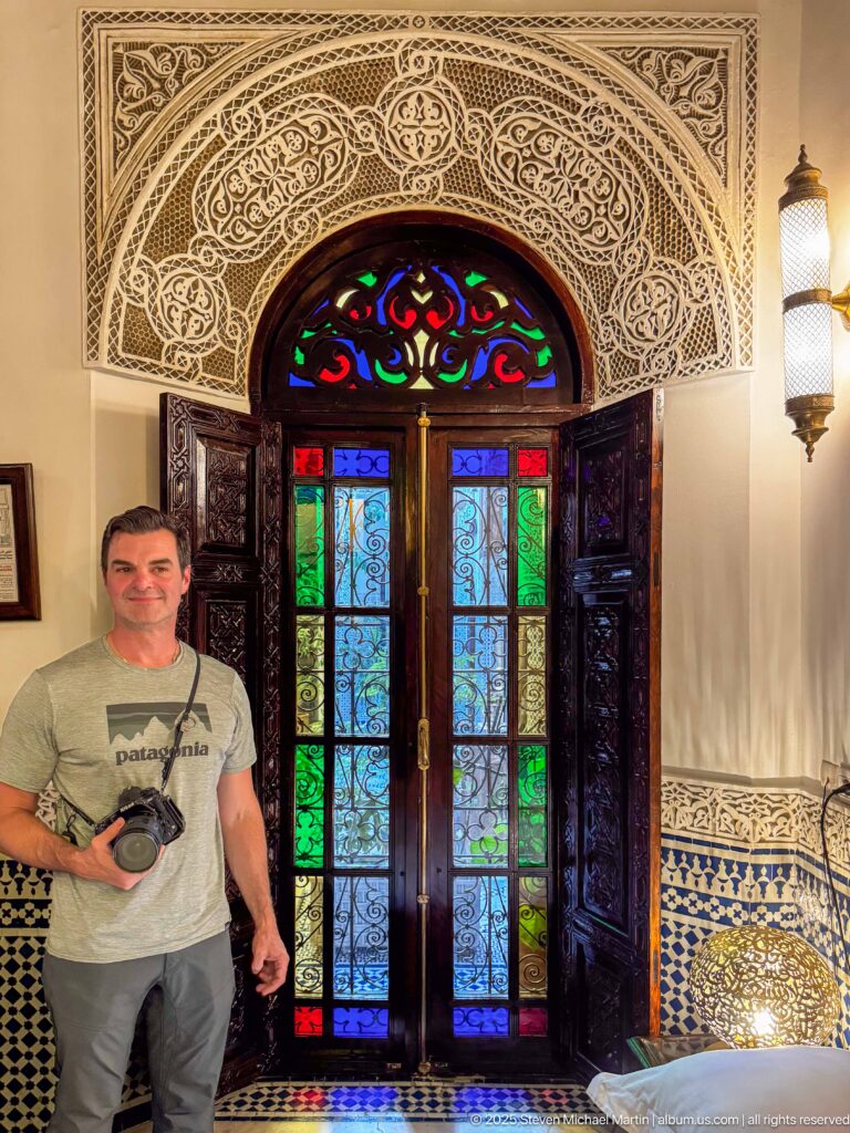 Man holding a camera beside tall carved doors with multicolored glass, ironwork, carved plaster, and zellij.