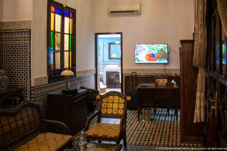Guest room with upholstered chairs, writing desk, television, zellij walls, patterned floor, and colored-glass doorway.