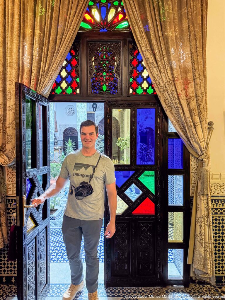 Man with camera standing in an open carved-wood doorway with multicolored glass panels, curtains, and tiled courtyard beyond.