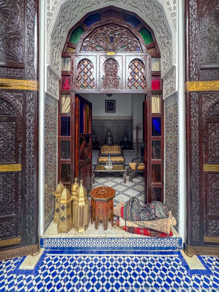Carved plaster and dark wood doorway with colored glass panels opening from a tiled courtyard into a furnished salon.