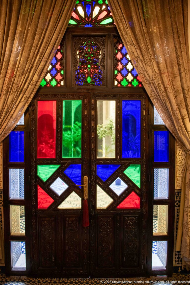 Symmetrical carved double doors with red, green, clear, and blue glass panels framed by draped curtains.