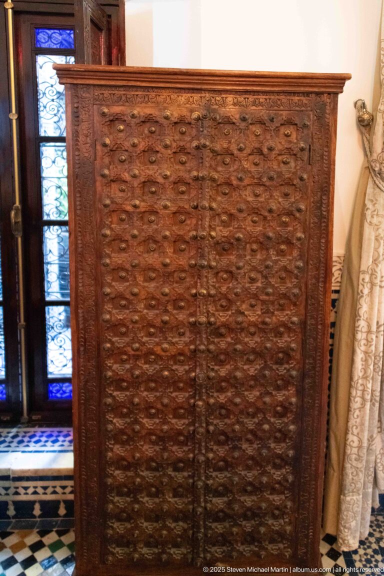 Tall brown wooden cabinet with two densely carved doors and small metal pulls beside colored glass and tiled wall.