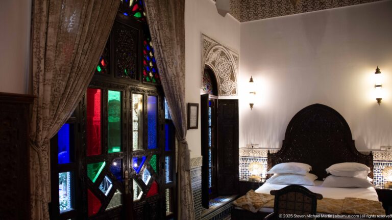Guest bedroom with carved headboard, white bedding, gold coverlet, colored-glass doors, curtains, zellij, and wall sconces.