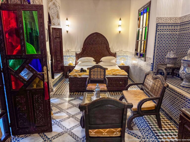 Wide guest bedroom with carved bed, patterned tile, chairs, small table, colored-glass doors, and ceramic vessels.