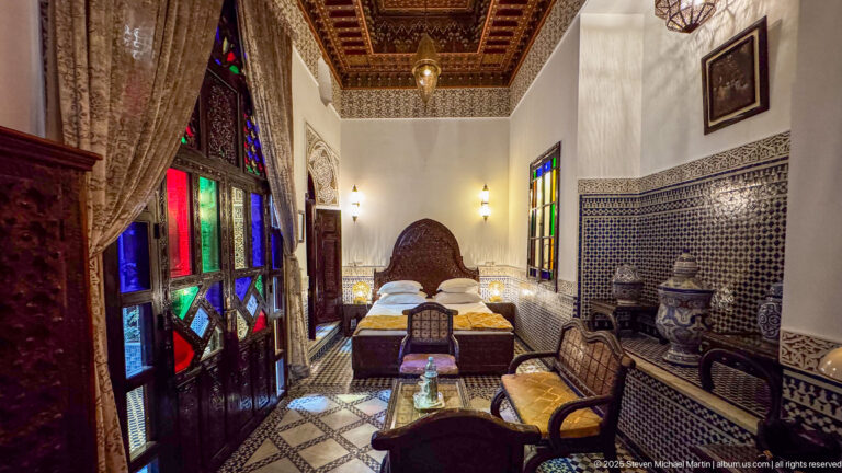 Long bedroom view with colored-glass doors, carved bed, zellij walls, patterned floor, seating, ceramics, and ornate wood ceiling.
