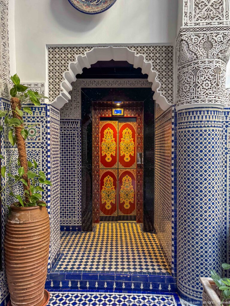 Scalloped arch lined with blue-and-white zellij framing red double doors with gold decoration and a large planter.