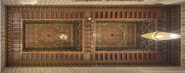 Upward view of an ornate coffered wooden ceiling with geometric panels, patterned borders, and suspended light fixtures.