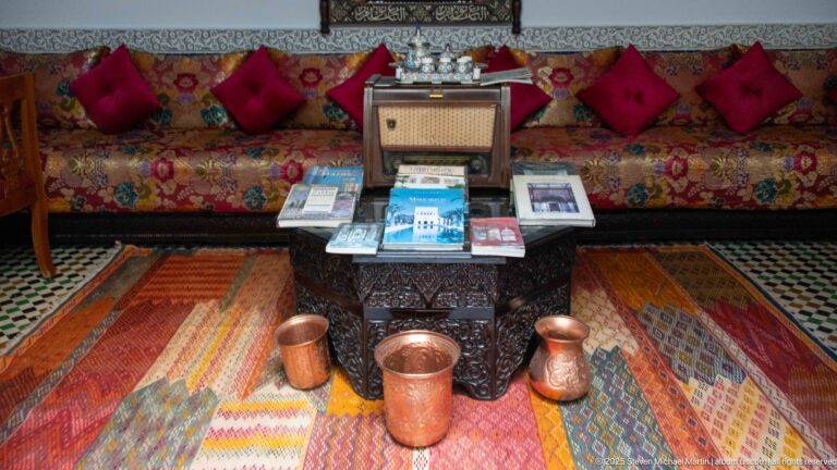 Low table with brochures and copper vessels before a patterned banquette with red cushions and colorful woven rugs.