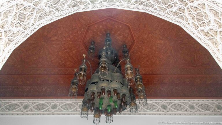 Large multi-tiered dark metal chandelier with green and clear glass hanging beneath a red ceiling and carved plaster arch.