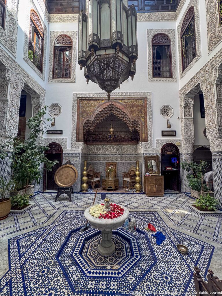 Riad courtyard with central fountain, blue-and-white tiled floor, carved plaster walls, brass vessels, plants, and hanging lantern.