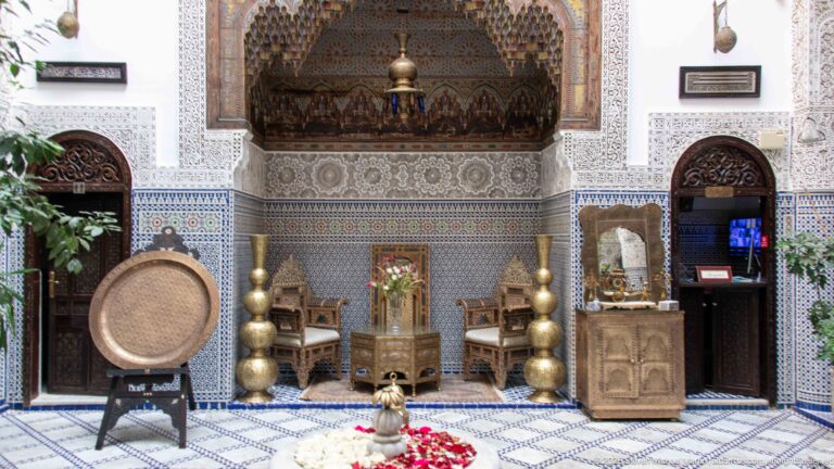 Ornate courtyard alcove with carved chairs, tall brass vessels, zellij, carved plaster, plants, and petal-filled fountain.