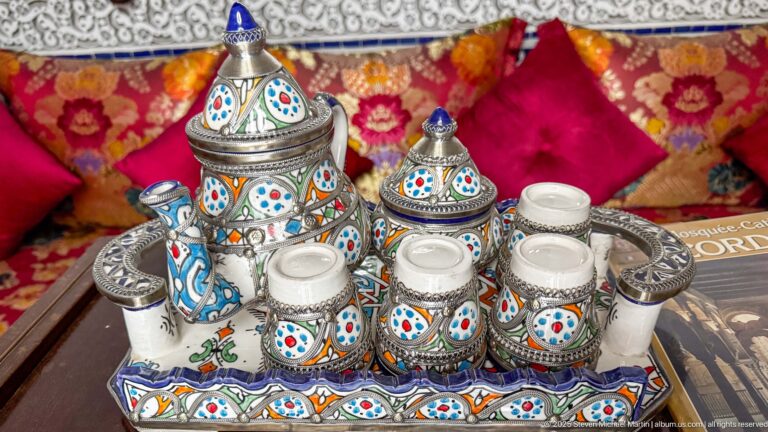 Colorfully painted lidded tea service and cups arranged on a tray in front of patterned red cushions.