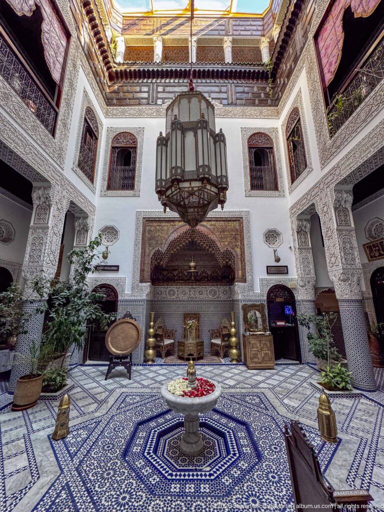 Symmetrical riad courtyard with tiled floor, central fountain, carved plaster walls, plants, and a large hanging lantern.
