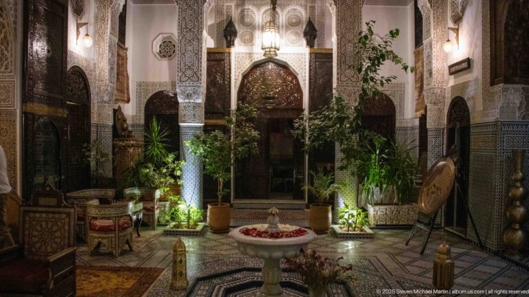 Night view of the riad courtyard with lit plants, carved plaster arches, low seating, patterned tiles, and a small central fountain.