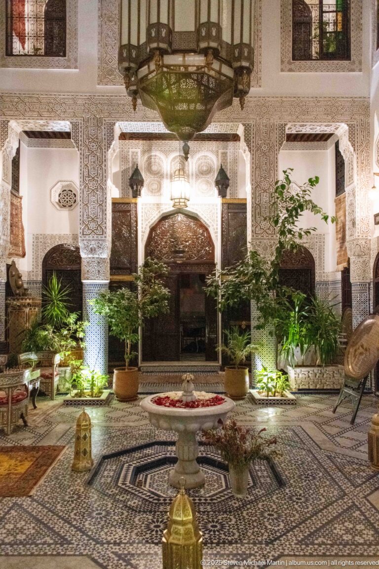 Illuminated riad courtyard at night with central fountain, patterned floor, plants, carved plaster, and hanging lantern.