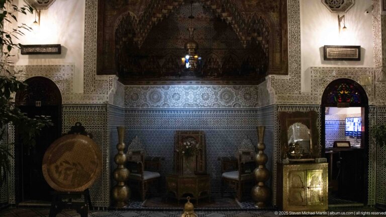 Dimly lit riad alcove with zellij walls, carved plaster, brass-toned vessels, cabinet, and large round tray.