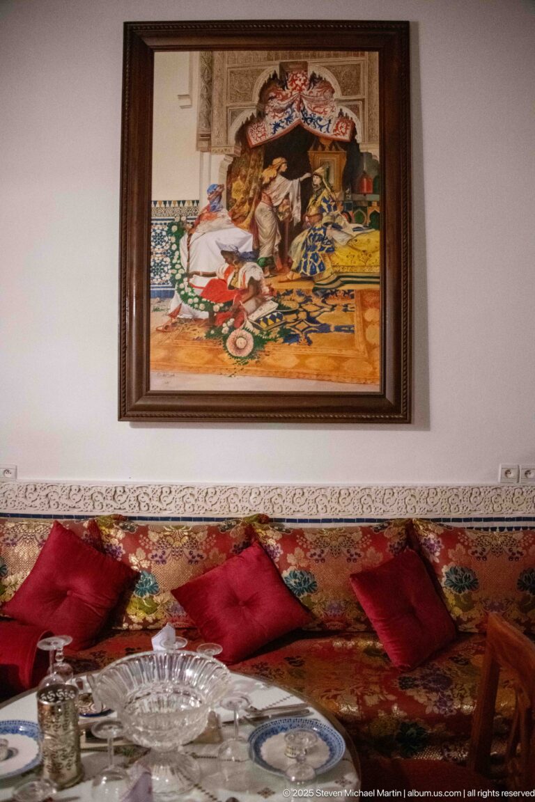Low Moroccan-style banquette with red patterned cushions, a set table, and framed figurative artwork above.