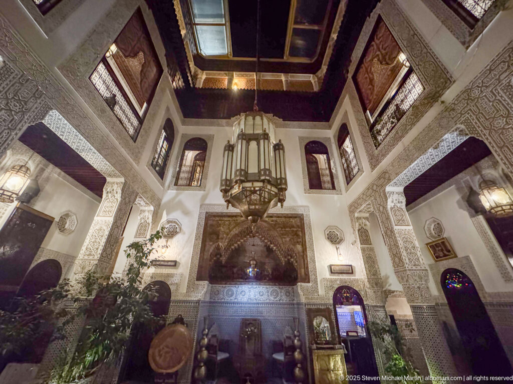 Upward view of a two-story riad courtyard with central lantern, carved plaster walls, upper windows, and roof opening.