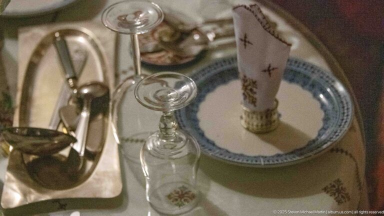 Dining place setting with blue-rimmed ceramic plate, folded embroidered napkin, stemmed glass, and polished metal service pieces.