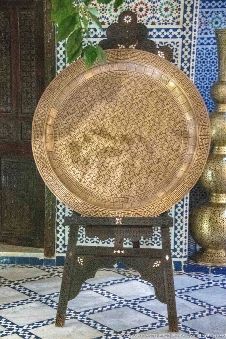 Large circular engraved brass-toned tray on a dark wooden folding stand against blue-and-white tiled walls.