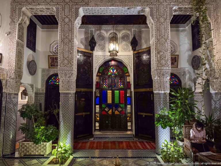 Arched dark-wood doorway with multicolored stained glass, carved plaster surround, zellij columns, and plants.