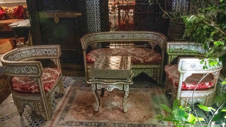 Low salon chairs, settee, and tables covered in geometric inlay with red patterned cushions and surrounding plants.