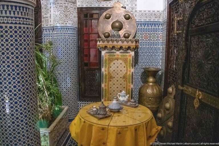 Brass-toned decorative wall feature with circular bosses behind a small round table and tea service in a tiled riad interior.