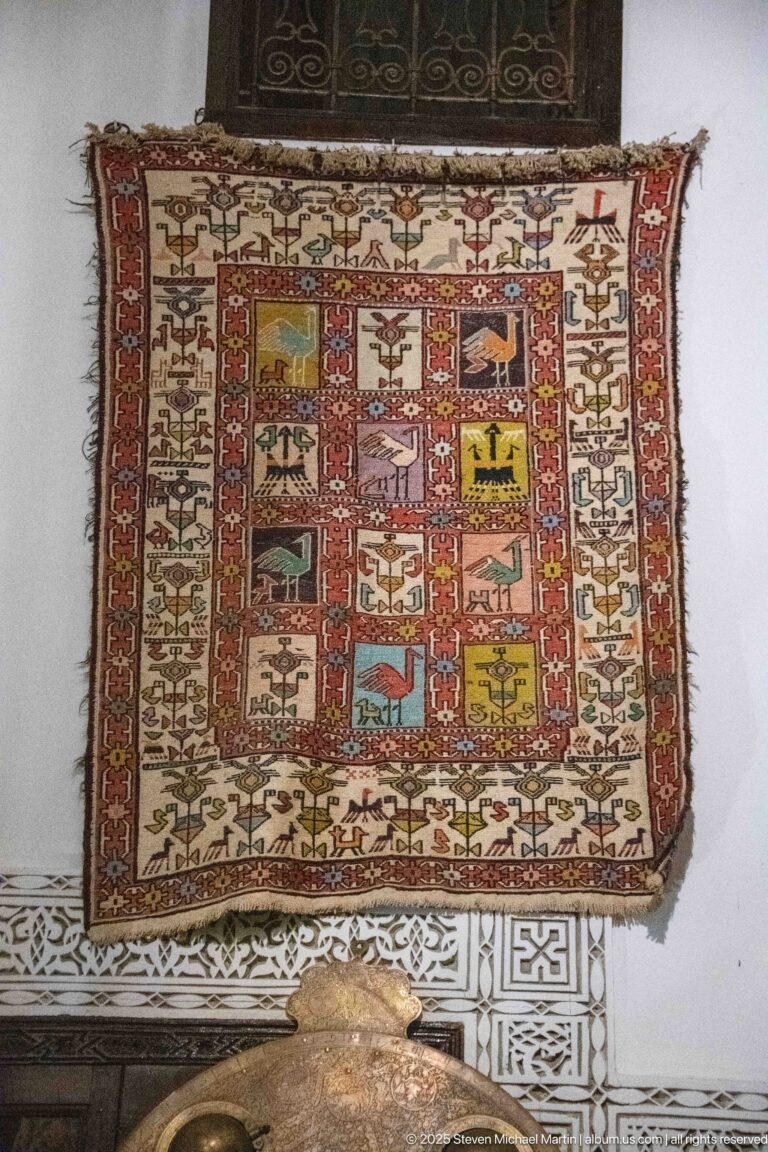 Rectangular wall-hung woven textile with colorful compartmented animal and human motifs above carved plaster trim.