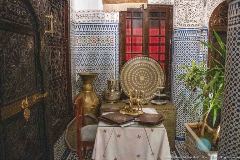 Brass-toned tray and vessels on a table beside carved wood, blue-and-white zellij, red glazing, and a potted plant.