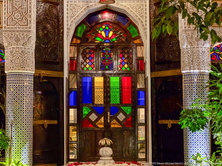 Frontal arched doorway with multicolored glazing, dark carved wood, carved plaster surround, tiled piers, and foliage.