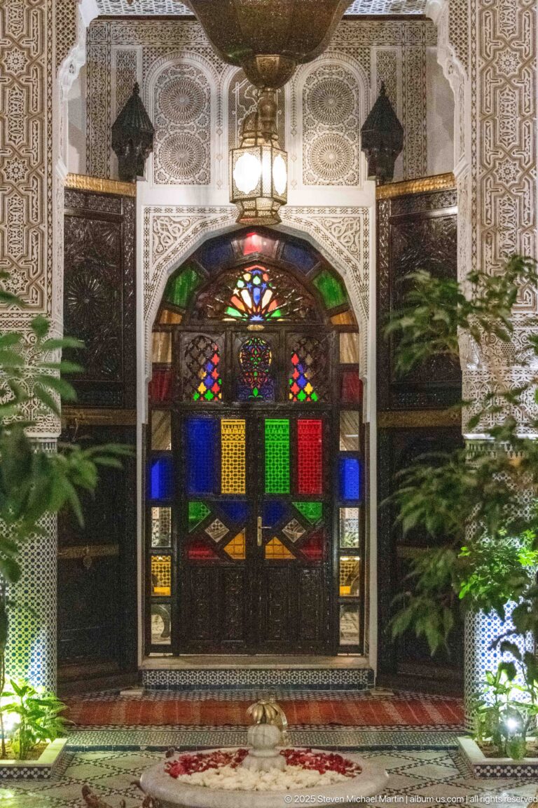 Multicolored glazed courtyard doorway aligned behind a low fountain, with carved plaster, tilework, and plants on both sides.