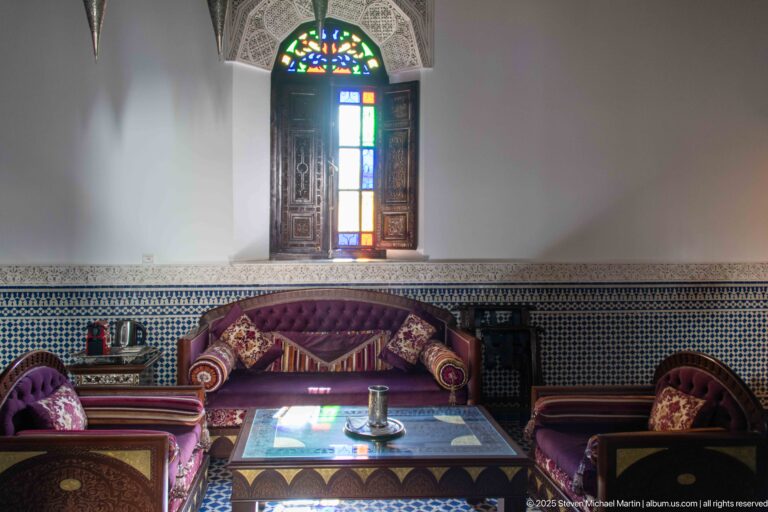 Low upholstered salon seating and table beneath a tall colored-glass window with zellij along the walls.