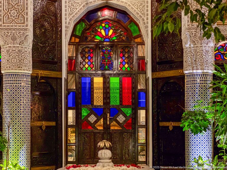 Close frontal view of multicolored glass panels above carved wooden doors between patterned plaster and zellij piers.