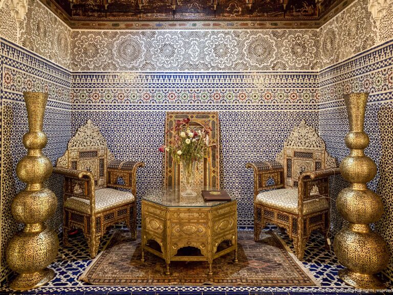 Symmetrical salon with two ornate armchairs, low table, central mirror and flowers, and tall pierced metal vessels against tiled walls.