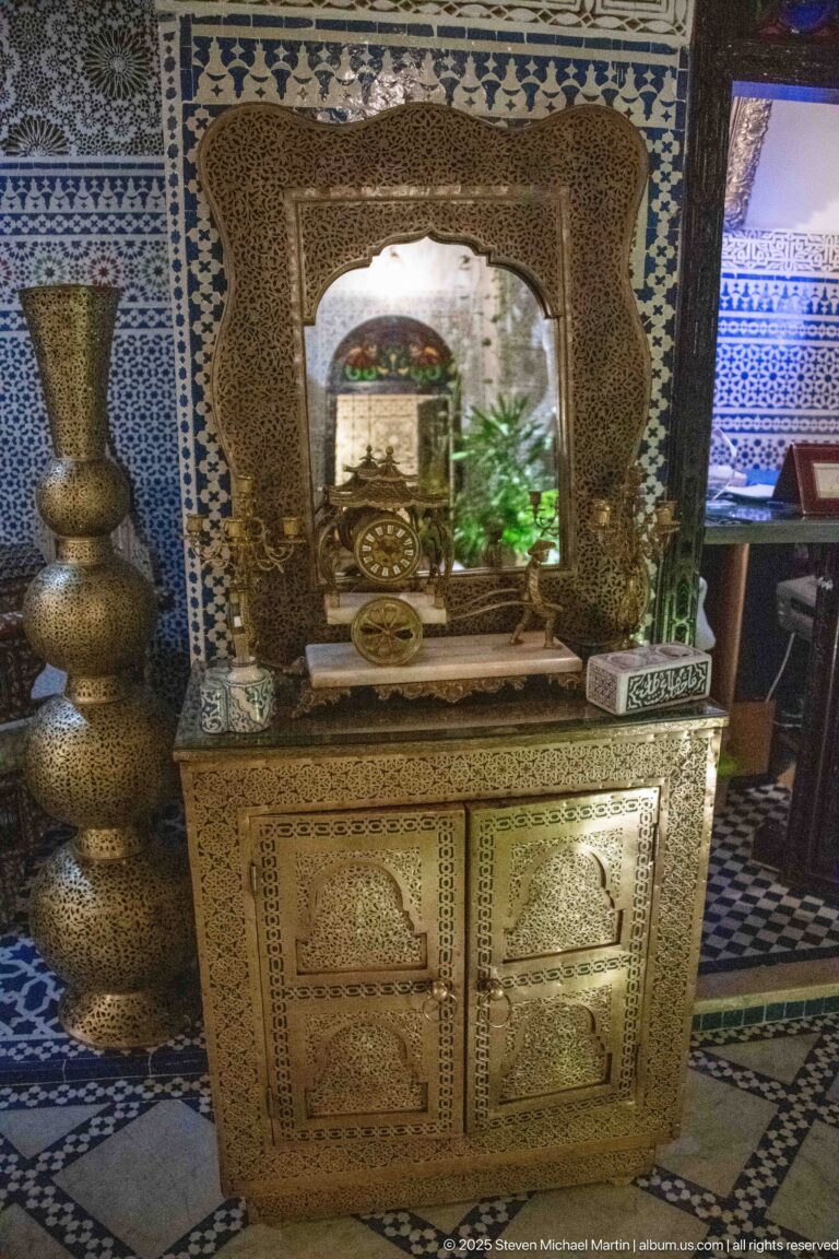 Decorative inlaid cabinet beneath a framed mirror, flanked by tall metal vessels against blue-and-white tiled walls.