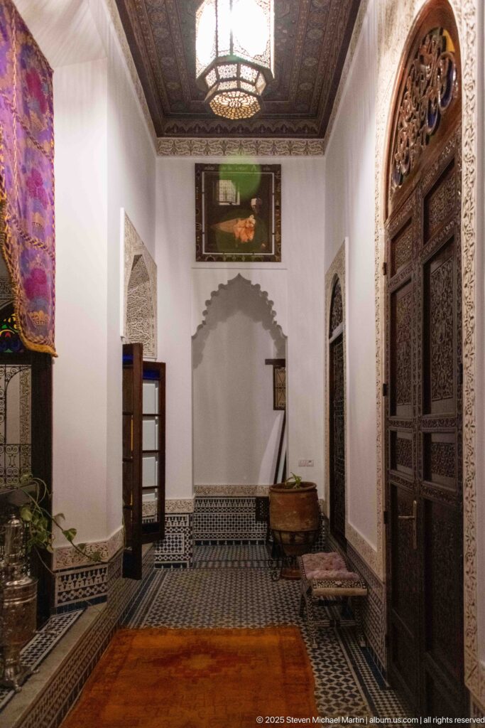 Riad interior passage with carved wooden doors, tiled lower walls, pale plaster ornament, bench, arched recess, and dark wood ceiling.