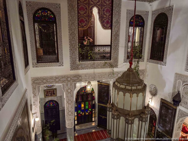 Upper-gallery view of the riad courtyard with a large foreground lantern and multicolored glazed doorway below.