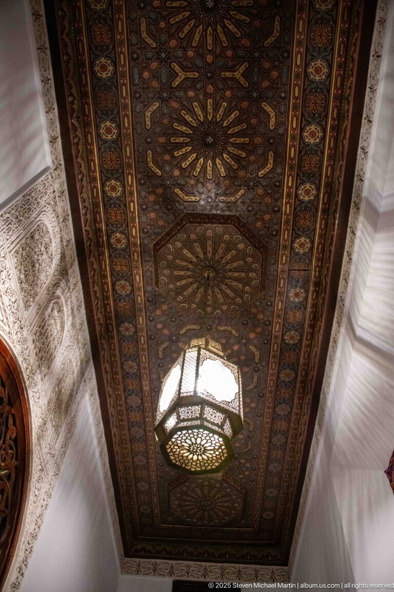Upward view of a dark geometric wooden ceiling with star-like medallions, painted borders, and a hanging lantern.