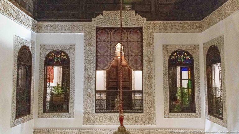 Upper riad wall with tall narrow windows, carved plaster surrounds, colored-glass arches, central wooden opening, and gallery rail.
