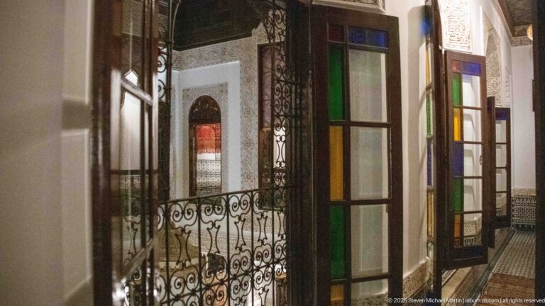 Upper gallery with tall wooden doors containing multicolored glass panels beside a scrolling wrought-iron balustrade.