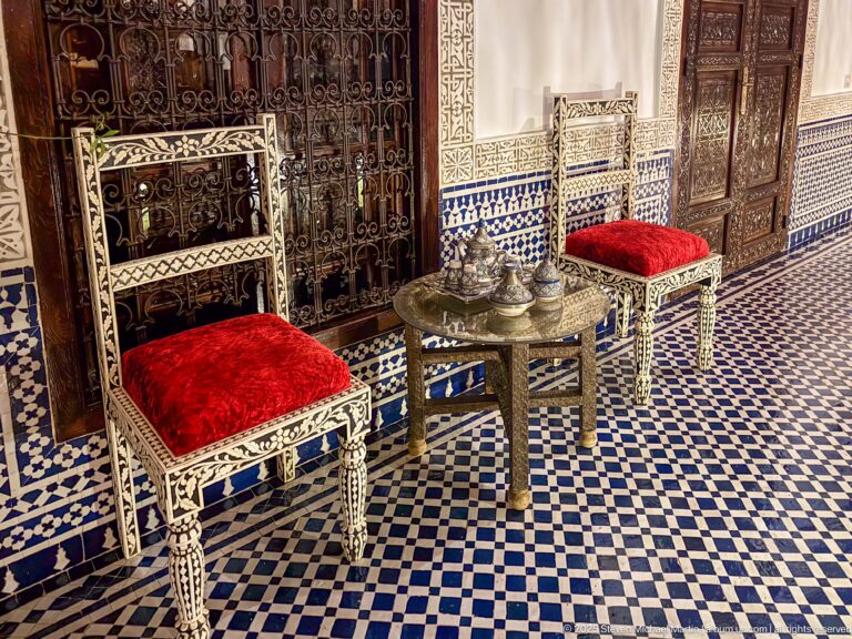 Two ornate high-backed chairs with red velvet seats beside a small round tea table on blue-and-white patterned tiles.