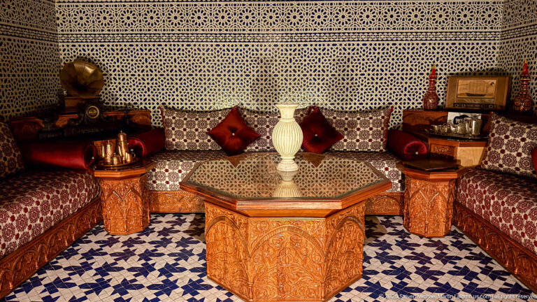 Moroccan-style salon with low patterned banquettes, carved wood tables, red cushions, white lamp or vase, and blue-and-white tiled floor.