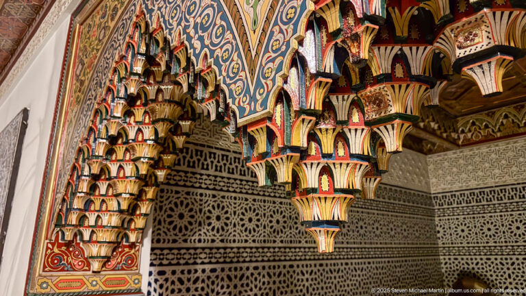 Close oblique view of clustered multicolored carved-wood brackets beneath a painted ceiling edge and above a geometric screen.