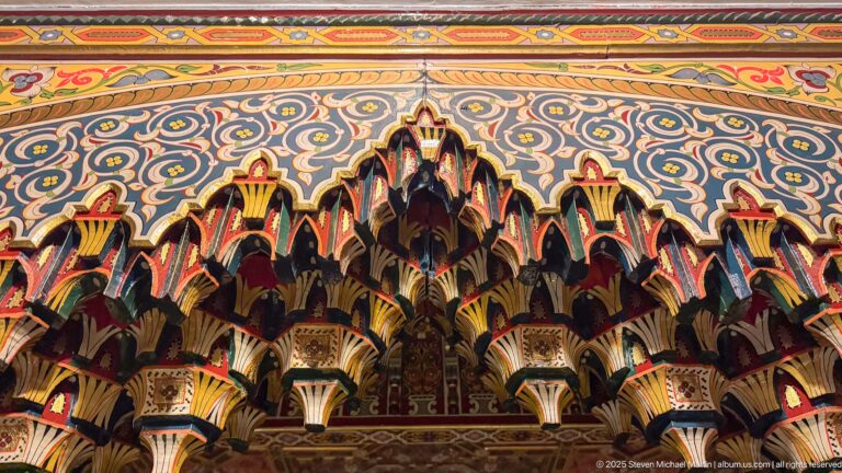 Frontal close-up of layered multicolored wooden brackets beneath scrolling painted ceiling decoration.