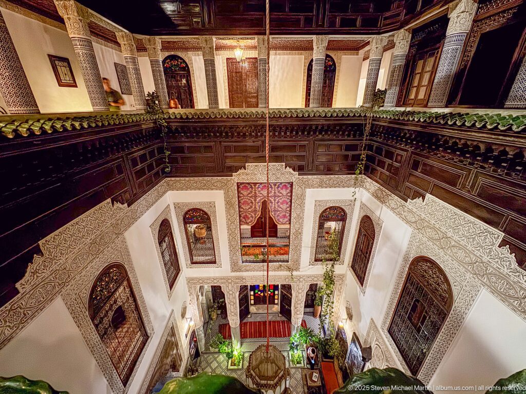 Wide upper-level view down into the riad courtyard with dark wooden galleries, carved plaster windows, plants, and suspended lantern.