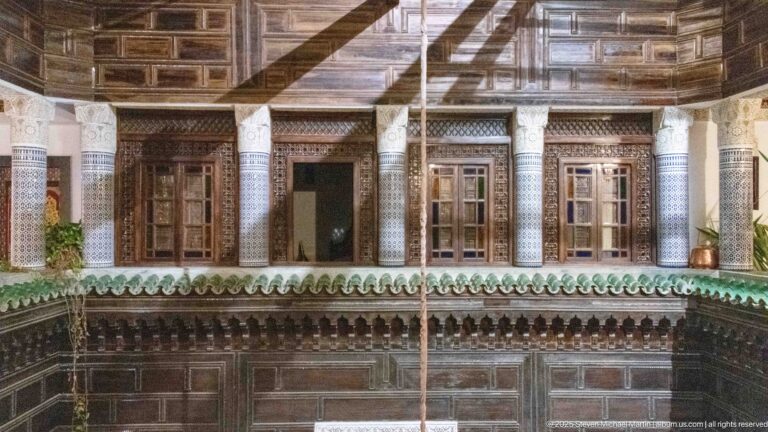 Straight-on view of a dark wooden upper gallery with carved columns, shuttered windows, paneled ceiling, and green-tiled eaves.